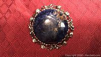 Top view of large blue stone as central cabochon with silver-tone floral petal metal frame around edge, on red textured surface.