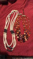 Photo showing two costume necklaces on a red surface, one faux pearl multi-strand with black spacers and one multi-strand amber and brown bead necklace.