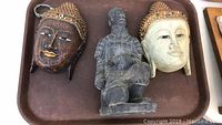 Pair of decorative masks and terracotta warrior statue shown together on a tray. Masks have bead decoration; statue is sitting in crouched position.