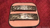 Pair of sterling silver etched hair barrettes placed on a pink and black display case, showing the front floral etched surface.