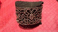 Front view of thick black hinged bracelet with black crystals and intricate openwork design on a black cushion with red textured background.