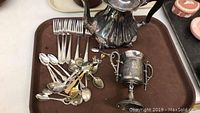 Overview of the silver teapot, double-handled chalice, and assortment of silver spoons and forks on a tray.