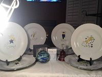 Photo of four large white dinner plates with silver rims, each featuring a different Christmas reindeer (Comet, Donner, Blitzen, Cupid), displayed on stands, along with a hand blown glass ornament and a glass bell.