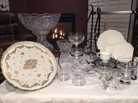Full lot of vintage Aynsley china, crystal glassware including pedestal punch bowl, vase, plates, and various glass drinkware on display on white cloth in front of fireplace.