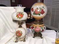 Photo showing front view of both vintage parlour hurricane lamps with floral painted glass and ornate metal bases, placed against white fabric background.