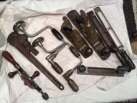 Photo showing assortment of antique hand tools including hand drills with red woodturning handles and wheel, a heavy Ridgid pipe wrench, several Stanley hand plane and rasp tools.