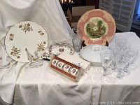 Full view of lot items on white draped cloth showing various plates, candy dish, napkin rings, glasses, casserole dish and figurine.