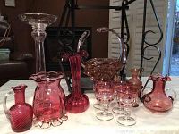 Full view of the lot with various pink cranberry and blown glass decorative items arranged on white cloth; includes vases, basket, creamer, goblets, and pitcher.