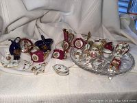Primary photo showing a collection of miniature porcelain pieces including blue and red watering cans, flower baskets, teapots, cups and decorative items arranged on white cloth