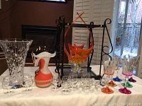 Table setup showing large clear crystal vase, white and orange decorative pitcher/vase, 8 small liquor glasses in front, multicolored stemmed wine glasses to right, and large orange footed hand-blown glass bowl in metal stand in background.