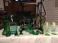 Full lot of green depression glass items arrayed on white cloth, including vases, glasses, horn of plenty vase, fruit bowl, and frog figurine.