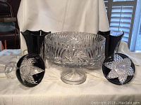 Front view of clear crystal pedestal centerpiece bowl flanked by two purple footed vases on white cloth background