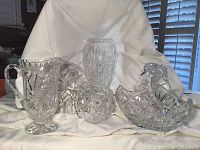 Photo showing the full set of crystal items: footed pitcher, vase, basket, and cake plate arranged on a white cloth backdrop.