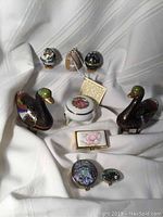 Photo showing the vintage cloisonne brass enamel items including egg-shaped ring holders, brass enamel ducks, porcelain and Mexican Alpaca pill boxes arranged on fabric.