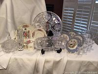 Full lot showing porcelain, crystal plates, bowls, glasses and serving pieces arranged together on white cloth backdrop with shutter window light.