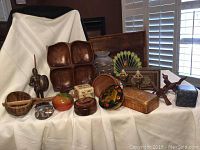 Full view of all items in the lot arranged on white cloth including teak serving ware, candy dish, folk art plate, brass shakers, knight figurine, trinket box and various wooden items