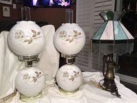 Pair of two lamps; one cast iron hurricane lamp with floral white globes, and one Tiffany style lamp with stained glass shade, shown on white cloth with dark background.