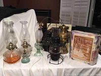 Seven vintage oil lanterns of various styles and sizes displayed, including a large brass lantern, a black small cage lantern, and multiple glass oil lamps with chimneys and bases in orange, clear, and green glass, plus a boxed Bordeaux twin oil lamp set with oil.