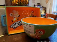 Side and inside view of large round ceramic bowl showing the orange interior with blue rim and part of the Rice Krispies characters and printed instructions on the exterior.