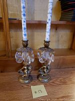 Full view of the pair of vintage brass candle holders each with decorative hanging crystal drops and tall white taper candle with blue floral design.