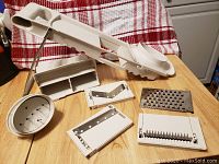 White Pampered Chef slicer and four blade attachments laid out on a wooden countertop with a red and white kitchen towel in the background.