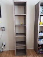 Full view of tall narrow book shelf with five shelves in grey woodgrain finish, standing on floor against wall.