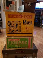 Three stacked boxes for Kodak Brownie 300 movie projector, Hanimex Electromatic 500 slide projector, and Bell & Howell case on top.