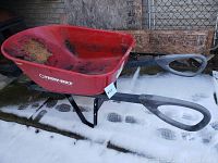 Red Troy-Bilt metal wheelbarrow with rust spots and wear on the tray surface, showing the entire wheelbarrow from the side including handles and support frame.