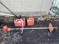 Full view of Stihl string trimmer laid next to two red gas cans on asphalt surface