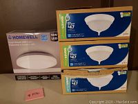 Four boxed flush mount LED light fixtures on a table against a brown wall. Three Globe Energy Saver 12-inch fixtures stacked on the right and one Homewell 16W LED fixture on the left.