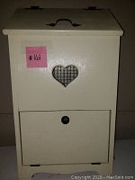 Front view of cream-colored wooden bin showing hinged top lid with black metal handle, heart-shaped wire mesh vent, and drop-down door with knob.