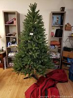 Full view of 6-foot green artificial Christmas tree assembled inside a room with shelves and various household items in the background, black plastic stand supporting the tree, red storage bag on the floor beside it.
