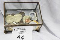 Brass and glass footed box containing the mixed collection of coins and tokens from above, showing the overall container and contents inside with top lid closed.