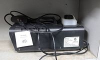 Side view of black rectangular fog machine with attached cord and wired remote placed on a shelf.