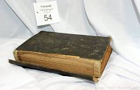 Overall view of the closed antique cookbook showing the black cover and worn spine with detached edges.