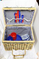 Open wicker picnic basket showing blue and white gingham lining, red and blue utensils inside lid, plastic dishes and cups, and plaid napkins inside basket.