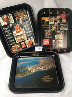 Three black metal Coca Cola collector trays laid out showing all fronts with distinct Coca Cola artwork and dates.