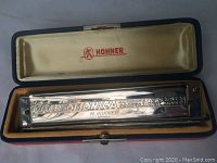 Open music case showing the silver chromatic octave professional harmonica with engraved model info and Hohner branding, inside cream and orange velvet-lined interior with red Hohner logo on lid.
