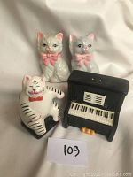 Two sets of cat-themed ceramic salt and pepper shakers including grey cats with pink bows and a cat playing the piano with matching piano shaker.