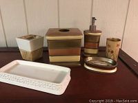 Photo of six bathroom accessory pieces on a dark surface, showing a tissue box holder, soap dish, ceramic cup, soap pump, toothbrush holder, and white porcelain tray.