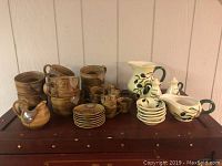 Full set of kitchen ceramics including brown soup bowls, espresso cups with saucers, and cream-colored olive oil dipping set and pitcher on a wooden surface