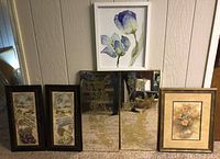Front view of 6 framed items. On top center, framed watercolor of two blue and white tulips in a simple white frame. Below is a pair of decorative mirrors with gold botanical leaf designs in dark frames. On left and right are four vertical framed pictures with vineyard landscapes, two on left in darker frames showing grapes and countryside scenery, one on right with floral arrangement in a gold-tone frame.