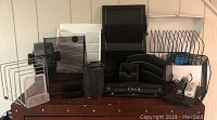 Overview of the lot showing various black and faux wood office desk supplies including file organizers, stackable organizers, mesh pen holder, stapler, and business card holder arranged on a wooden desk.