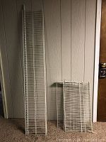 Two white wire shelving units, one tall and narrow, the other shorter, leaning against a wall.