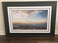 Framed print showing the sunset over ocean rocks in black frame with glass, positioned on dark wood surface.