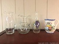 Four pitchers on a brown surface: three clear glass pitchers of different shapes and one ceramic colorful floral pitcher.