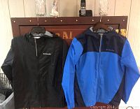 Two youth large Columbia windbreakers hanging on hangers, one black and gray and one blue and navy.