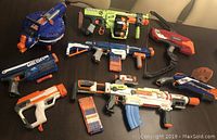Photo showing the full lot of 8 Nerf guns arranged on a dark surface, displaying various models and colors, some with loaded magazines and darts.