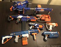 Top view of eight assorted Nerf guns laid out on a dark surface showing variety of sizes and styles with orange darts loaded.