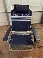 Front view of the Nautica folding chair showing the navy canvas seat and backrest with stripes and logo, metal frame, and armrests.
